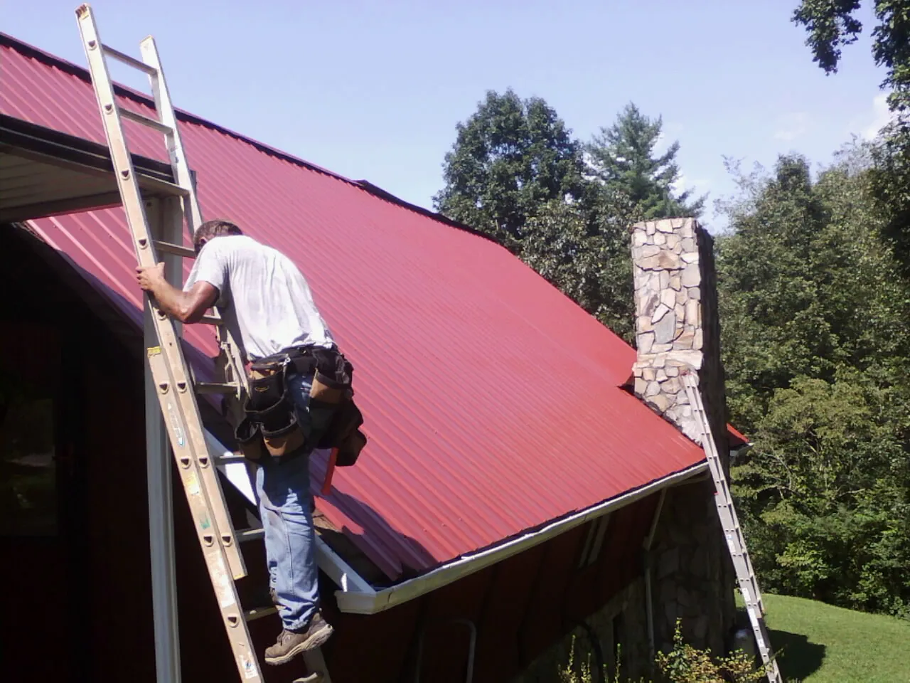 Roofer on a ladder beside a red metal roof and stone chimney — Soffit & Fascia Repair by Roofing Brothers in Maury